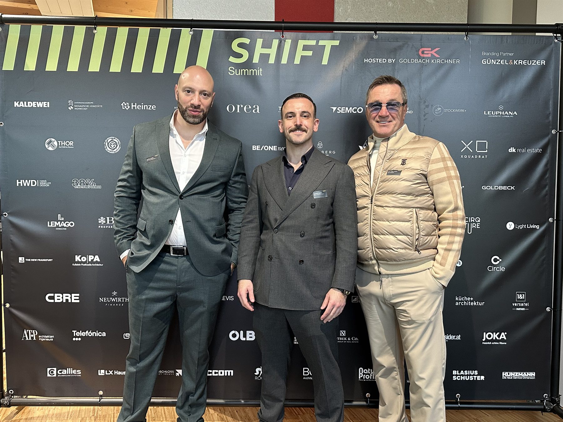 Daniel Talsik, Cengiz Seker und Ralf Kollinger vor der Pressewand des SHIFT Summit 2026 bei Goldbach Kirchner
